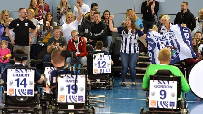Five people in powerchairs taking part in a powerchair football match in a sports hall while spectors cheer animatedly in the background