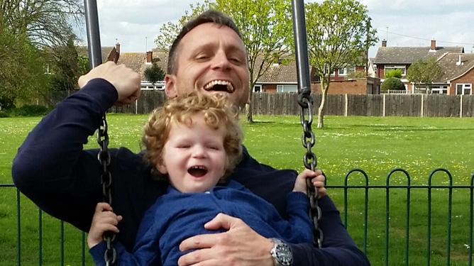 A toddler diagnosed with Congenital Muscular Dystrophy laughing with his dad while the sit together on a swing.
