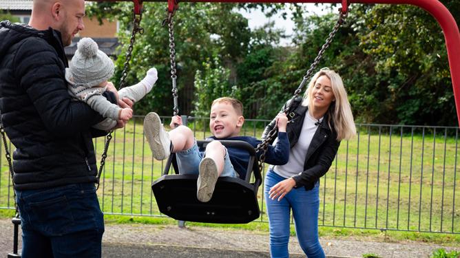 A father holds a young baby while a mother pushes her young son, who has Duchenne muscular dystrophy, on a swing.