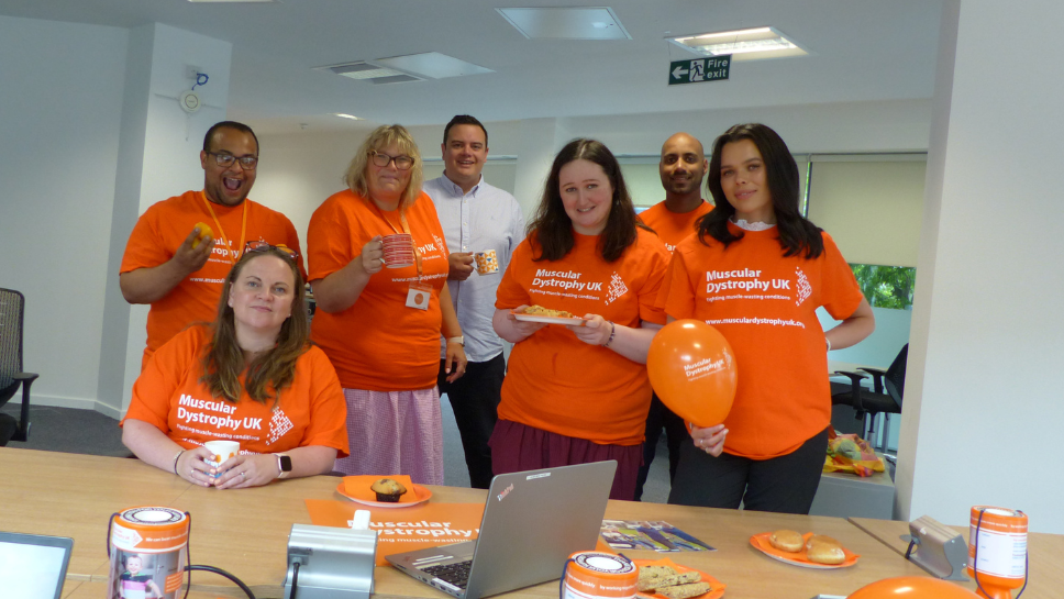 Image of 7 volunteers in their MDUK Orange T-shirts holding cakes, balloons and in front of a table of fundraising materials