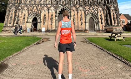 A man standing in front of Lichfield cathedral. His back is to the camera. He wears a Muscular Dystrophy UK running vest. It is bright orange and teal.