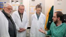 A seated researcher in a green lab coat in discussion with three colleagues wearing white lab coats.