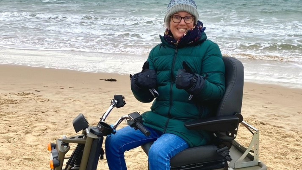 Louise is sat on a mobility scooter on the beach, smiling with her thumbs up. She's wearing a green puffer jacket and a woolly hat